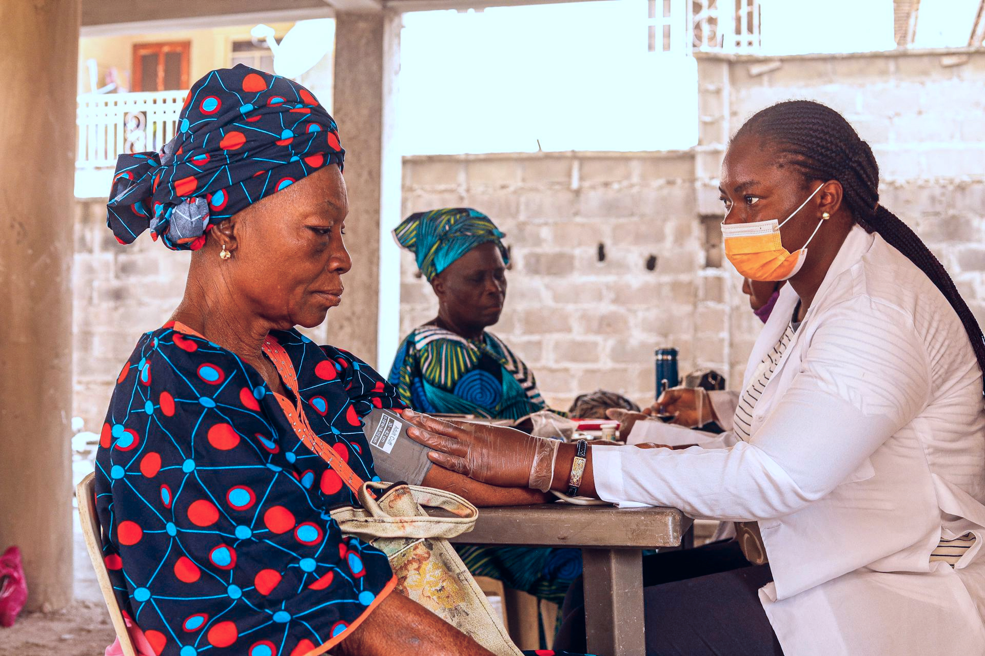 A healthcare worker wearing a white coat, gloves, and a face mask checks the blood pressure of an older woman dressed in a colorful outfit and headwrap. A healthcare worker wearing a white coat, gloves, and a face mask checks the blood pressure of an older woman dressed in a colorful outfit and headwrap.
