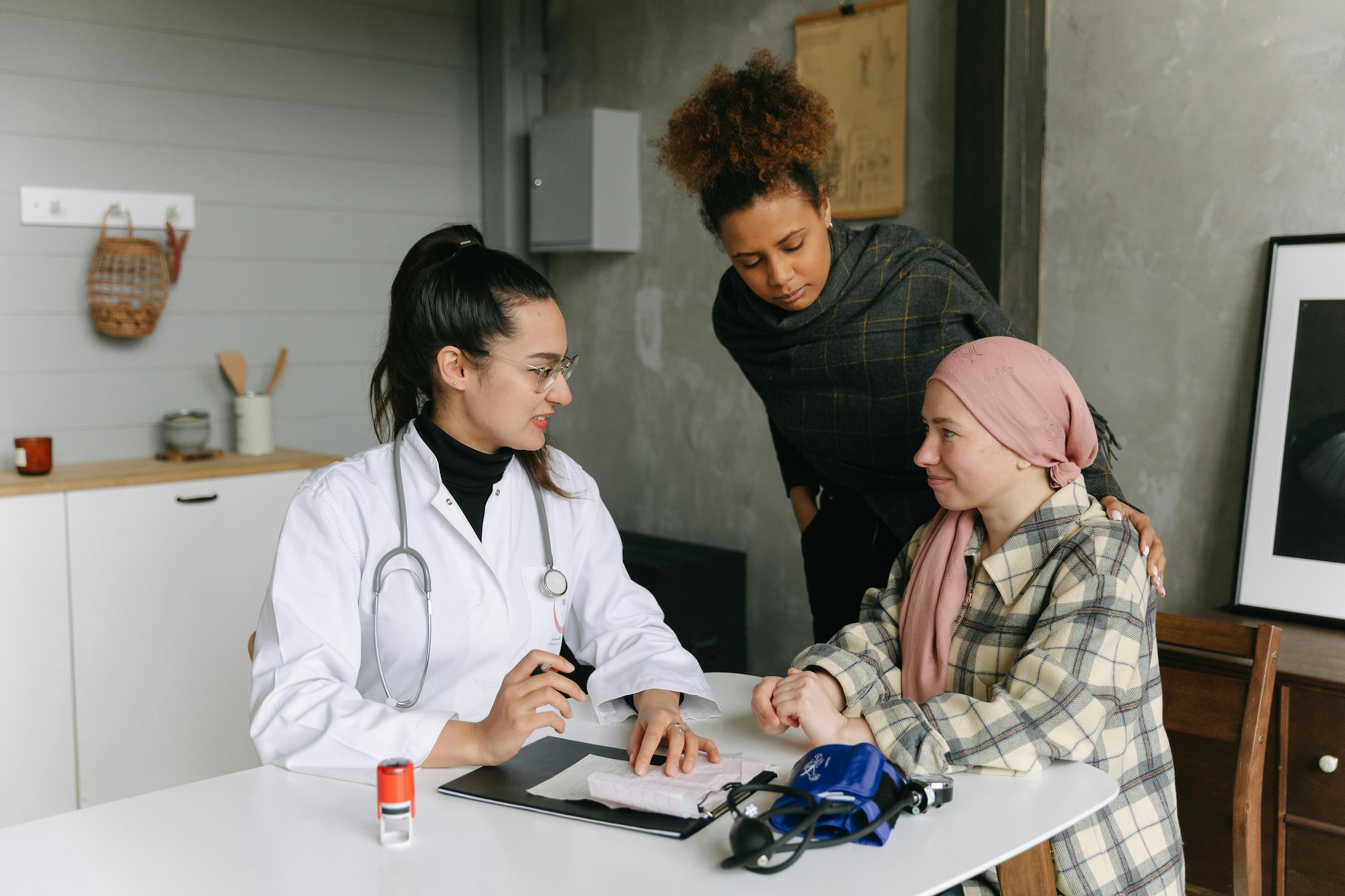 A female doctor in a white coat and stethoscope sits at a table talking with a patient wearing a headscarf and plaid jacket. Another woman stands beside the patient, offering support. A female doctor in a white coat and stethoscope sits at a table talking with a patient wearing a headscarf and plaid jacket. Another woman stands beside the patient, offering support.
