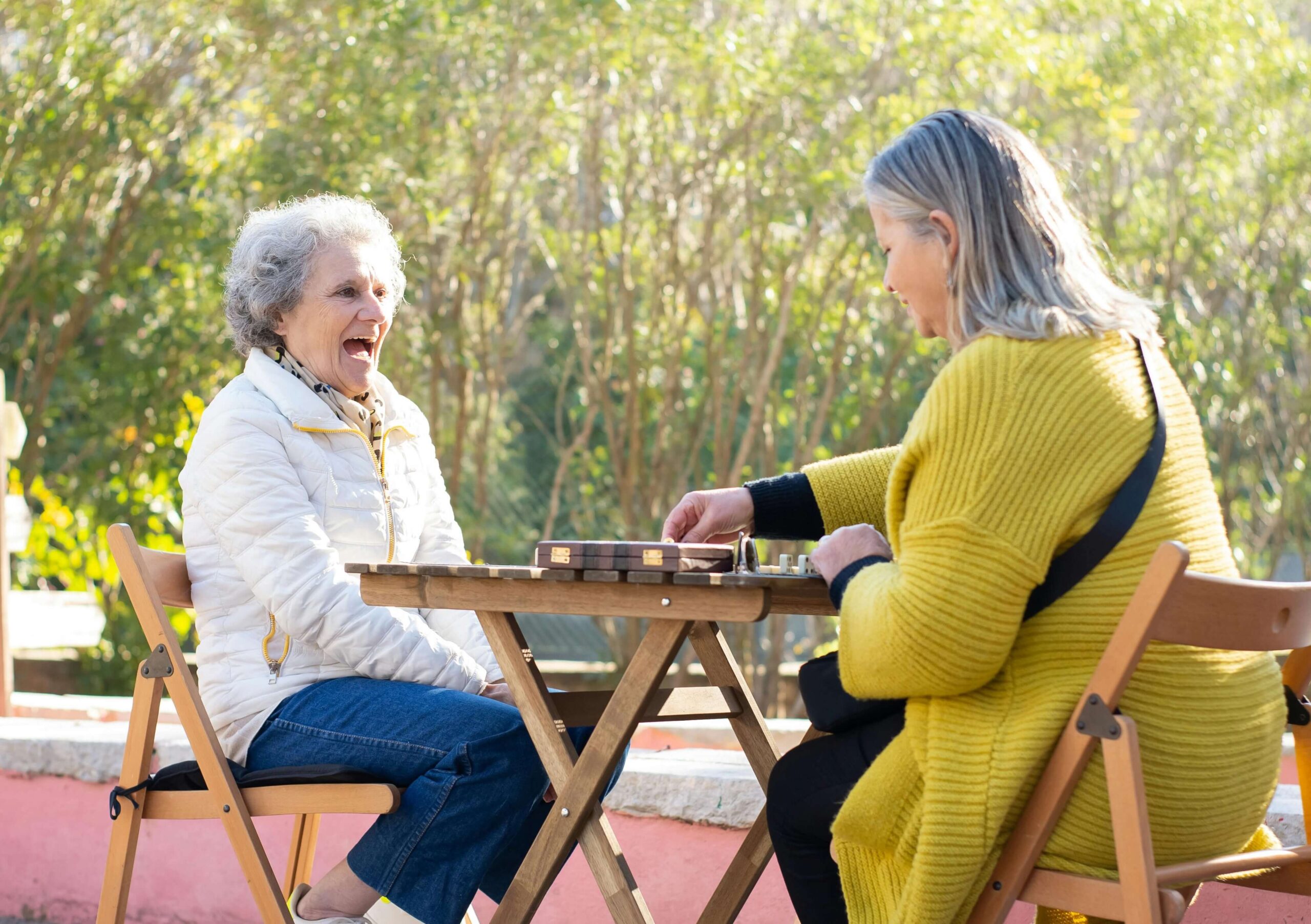 Two ladies sit happily across from each other at an outdoor table playing dominoes. Two ladies sit happily across from each other at an outdoor table playing dominoes.